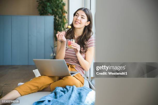 young woman sitting at the window at home with laptop having a snack - fruit salad stock pictures, royalty-free photos & images
