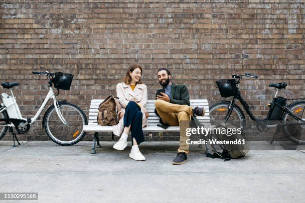 happy couple sitting on a bench next to e-bikes sharing cell phone - banco asiento fotografías e imágenes de stock