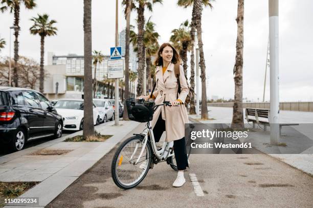 smiling woman with e-bike on a promenade - trench coat stock pictures, royalty-free photos & images