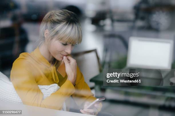 blond businesswoman sitting at window, doing a paymant with smartphone and creditcard - red blouse fotografías e imágenes de stock