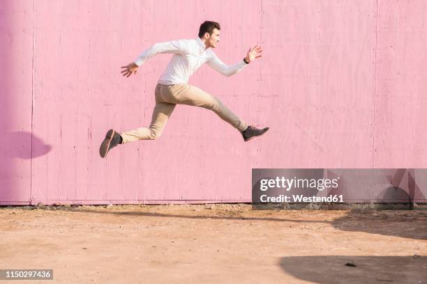 exuberant young man jumping in front of pink wall - man jumping stock pictures, royalty-free photos & images