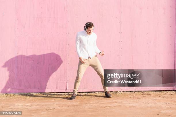 exuberant young man listening to music in front of pink wall - rock music stock-fotos und bilder