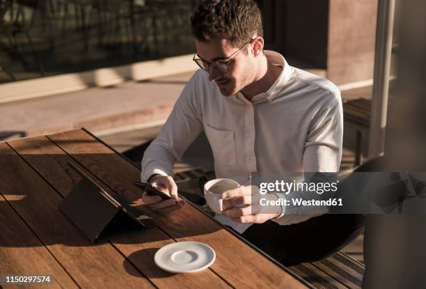 young man using cell phone and tablet at outdoor cafe - terrasse-de-café photos et images de collection