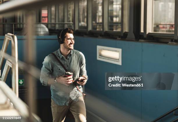 smiling young man with headphones, cell phone and takeaway coffee walking at the station - young man listening to music on smart phone outdoors stockfoto's en -beelden