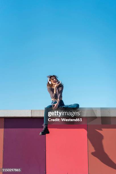 young woman sitting on a wall listening music with headphones on her smartphone - barcelona free stock-fotos und bilder