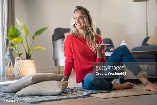 smiling woman sitting on the floor at home holding cell phone - donne bionde scalze foto e immagini stock