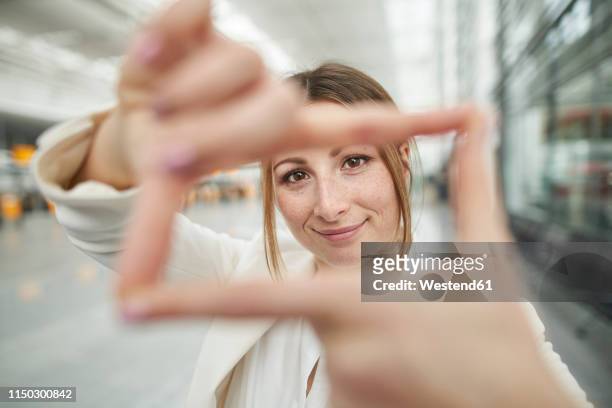 portrait of smiling young businesswoman doing finger frame at the airport - fingerrahmen stock-fotos und bilder