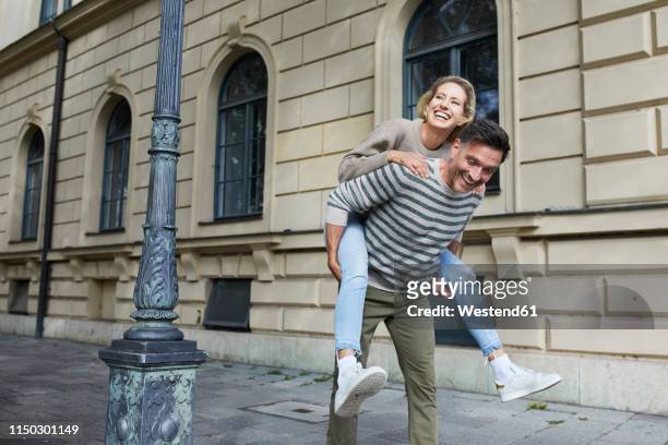happy man giving woman piggyback ride on pavement in the city - piggyback stock pictures, royalty-free photos & images