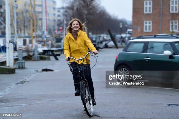 denmark, copenhagen, happy woman riding bicycle on waterfront promenade in rainy weather - blond woman city lifestyle stock-fotos und bilder