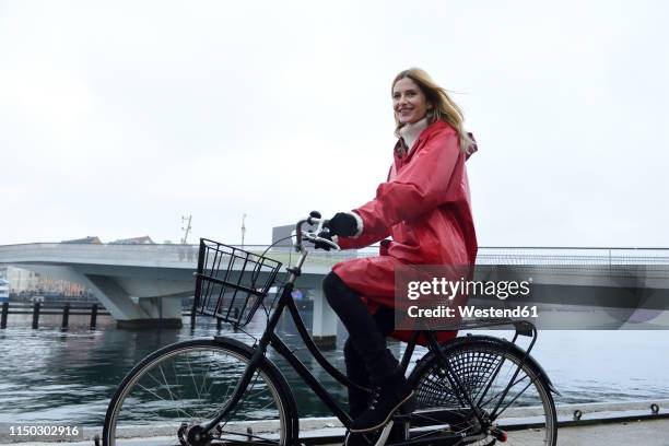 denmark, copenhagen, happy woman riding bicycle at the waterfront in rainy weather - winter denmark stock pictures, royalty-free photos & images