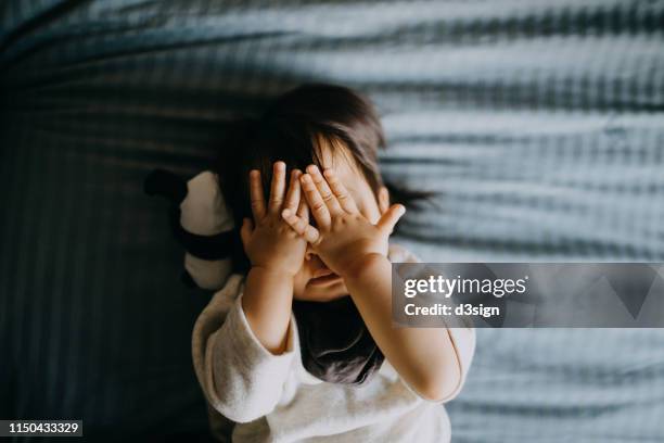 portrait of cute little asian toddler girl covering her face playing peekaboo while lying on bed with panda soft toy lying next to her - kids sleep in bed stock-fotos und bilder