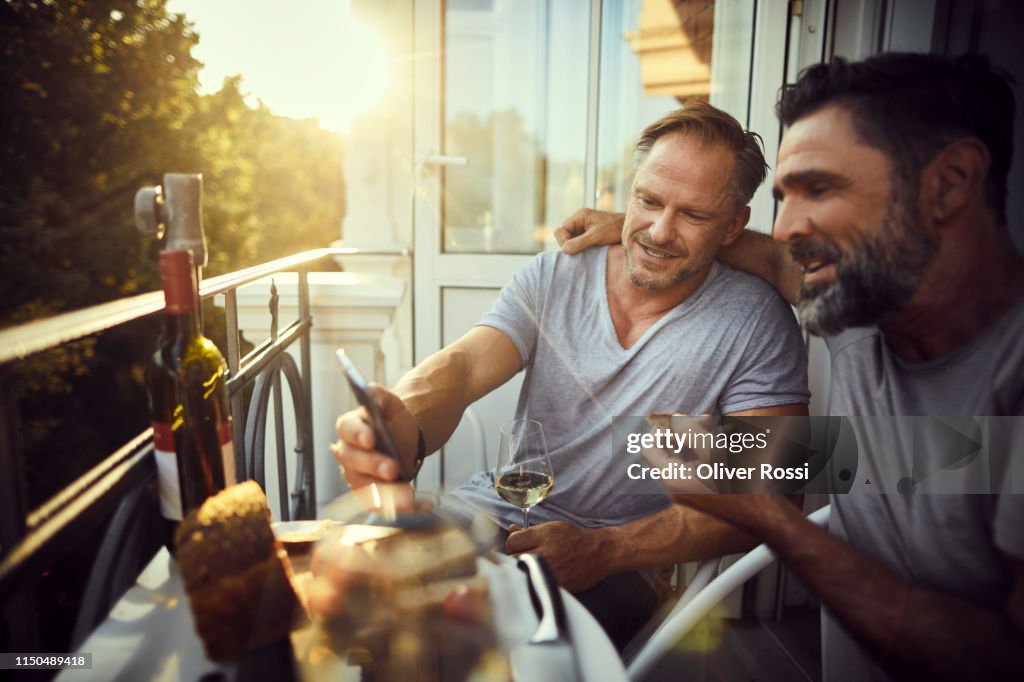 Two mature male friends with cell phone having dinner on balcony