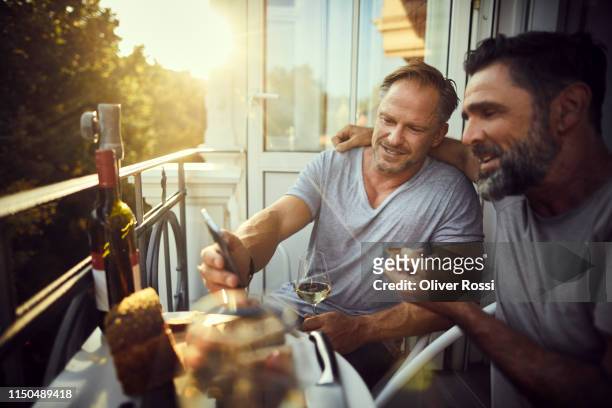 Two mature male friends with cell phone having dinner on balcony