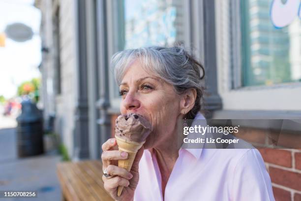 senior woman sitting on sidewalk eating chocolate ice cream cone - senior adult eating stock pictures, royalty-free photos & images