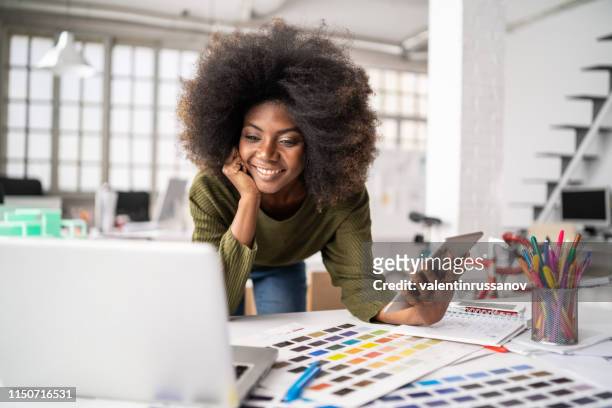 stilista africano che lavora in studio e utilizza laptop e smartphone - inserzione pubblicitaria foto e immagini stock