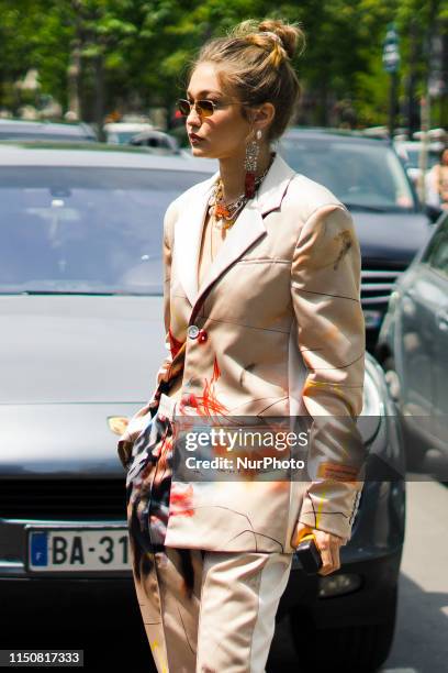 Gigi Hadid wears sunglasses, earrings, necklaces, a Heron Preston lustrous beige pantsuit with colorful paintings print, grey padded heeled mules, is...