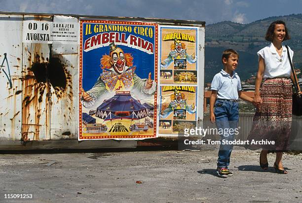 Signs Of The Times - A mother and her son walk past a poster advertising a circus carries the smiling face of a clown on a shell damaged wall during...