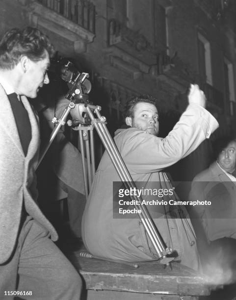 American film director Orson Welles, wearing a trench, directing on the set of 'Othello's movie, a tripod behind him and some members of the crew,...