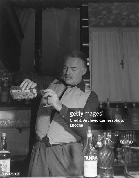 American writer Ernest Hemingway wearing a hunter waistcoat, standing behind a bar counter and pouring gin from a bottle of Gordon's, other alcoholic...