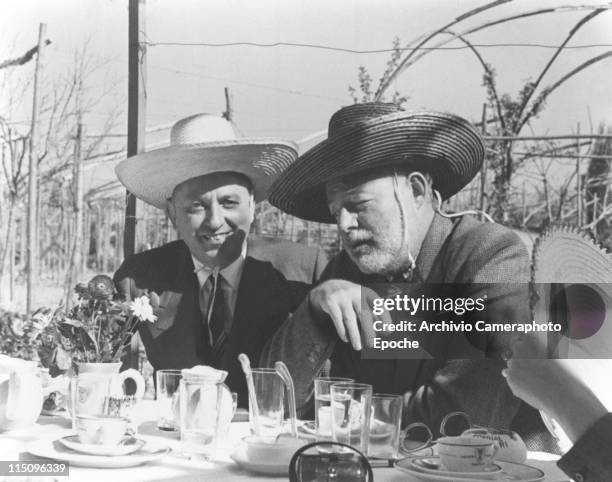 American writer Ernest Hemingway, sitting at a table with Giuseppe Cipriani, both wearing a sombrero straw hat, glasses, cups and flowers on the...