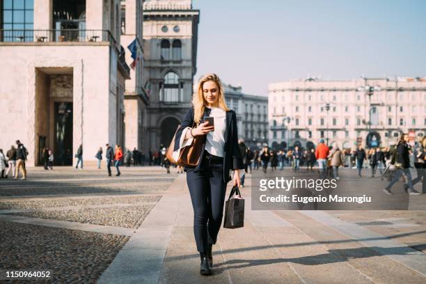 young female tourist with shopping bags strolling and looking at smartphone in city square, milan, italy - milan italy stockfoto's en -beelden