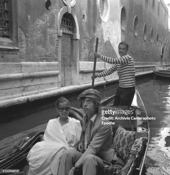British singer Mick Jagger, wearing a suit, sitting next to Bianca Jagger wearing a white poncho and sunglasses, in a gondola, with the gondolier...