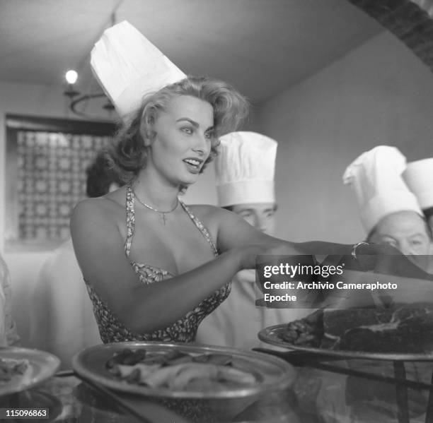 Italian actress Sophia Loren, wearing a floral dress, a chef's hat and a crucifix necklace, cutting slices of ham with a big knife, surrounded by...