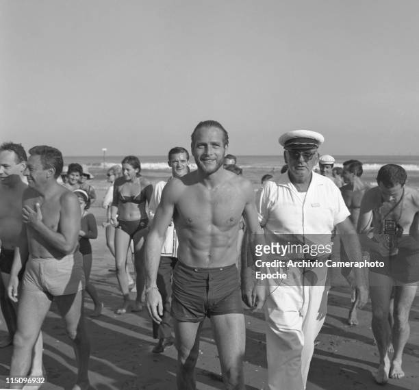 American actor Paul Newman, wearing a swimming suit, walking on the Lido seashore surrounded by the crowd, a man wearing a sailor cap and another one...