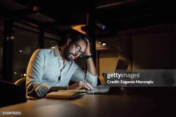 tired businessman in office at night typing on laptop - overworked man stock pictures, royalty-free photos & images