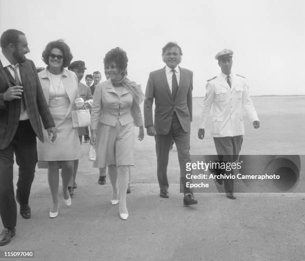 American actress Elizabeth Taylor, wearing a tailleur and a foulard, holding a handbag, walking next to her husband Richard Burton, many people...