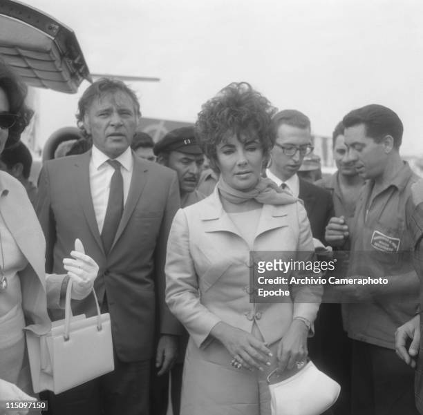 American actress Elizabeth Taylor, wearing a tailleur and a foulard, holding a handbag, in a close-up, next to her husband Richard Burton, many...