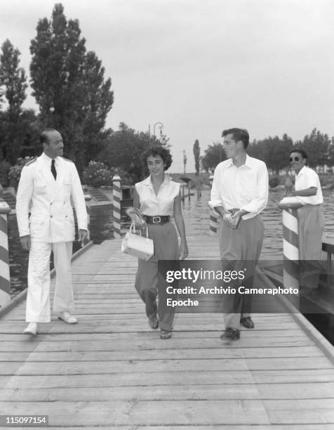 American actress Elizabeth Taylor, wearing a sleeveless shirt and wide trousers, holding a newspaper and a handbag, walking next to her first husband...