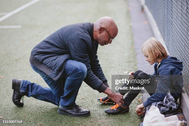 full length side view of grandfather helping grandson wearing soccer shoe on playing field - boy tying shoes stock-fotos und bilder