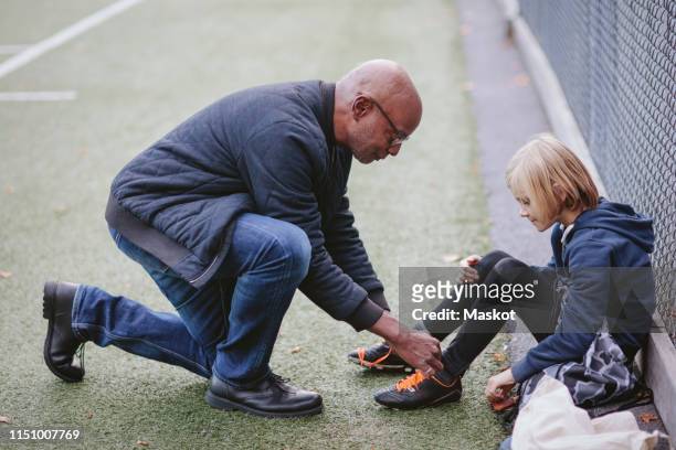 full length side view of grandfather helping grandson wearing soccer shoe on playing field - tiersport stock pictures, royalty-free photos & images