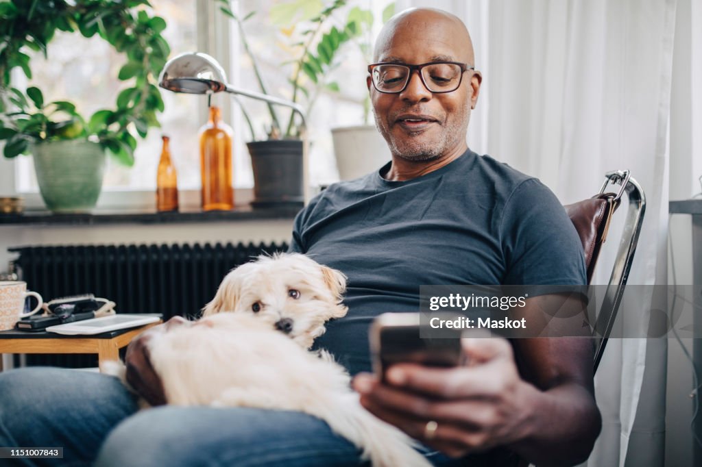 Smiling retired senior male using smart phone while sitting with dog in room at home
