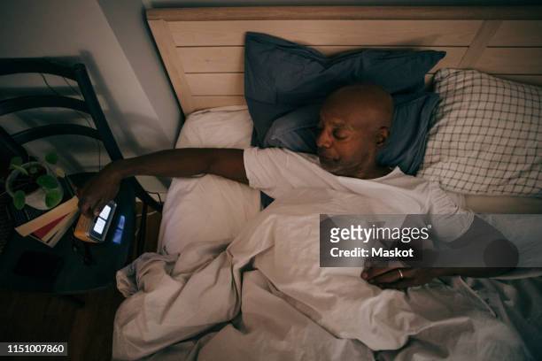 high angle view of retired senior man turning off alarm clock by bed at home - black man sleeping in bed - fotografias e filmes do acervo