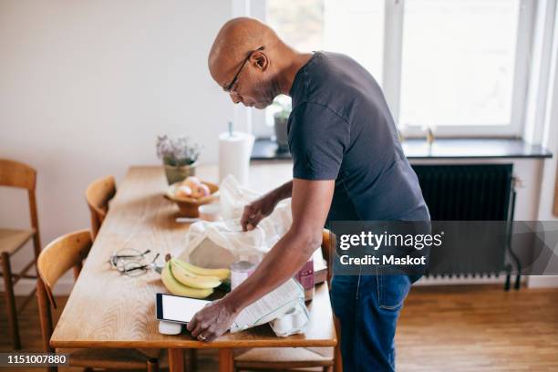 side view of retired senior man checking shopping list on digital tablet while removing groceries from bag in kitchen at - boodschappenlijst stockfoto's en -beelden