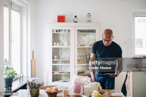 elderly man checking shopping list on digital tablet while removing groceries from bag in kitchen at home - old person kitchen food stock-fotos und bilder