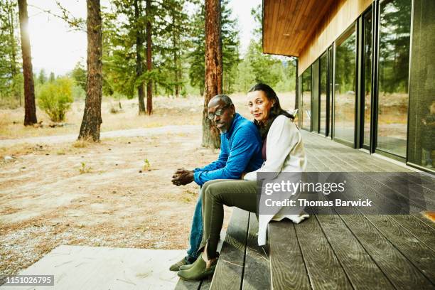 senior couple sitting together on porch of cabin in woods - ritratto coppia mista foto e immagini stock