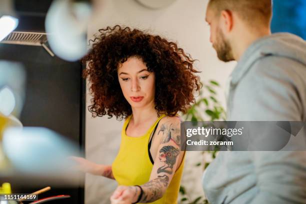 young tattooed woman with curly hair by the kitchen counter with roommate - lady cooking confused imagens e fotografias de stock