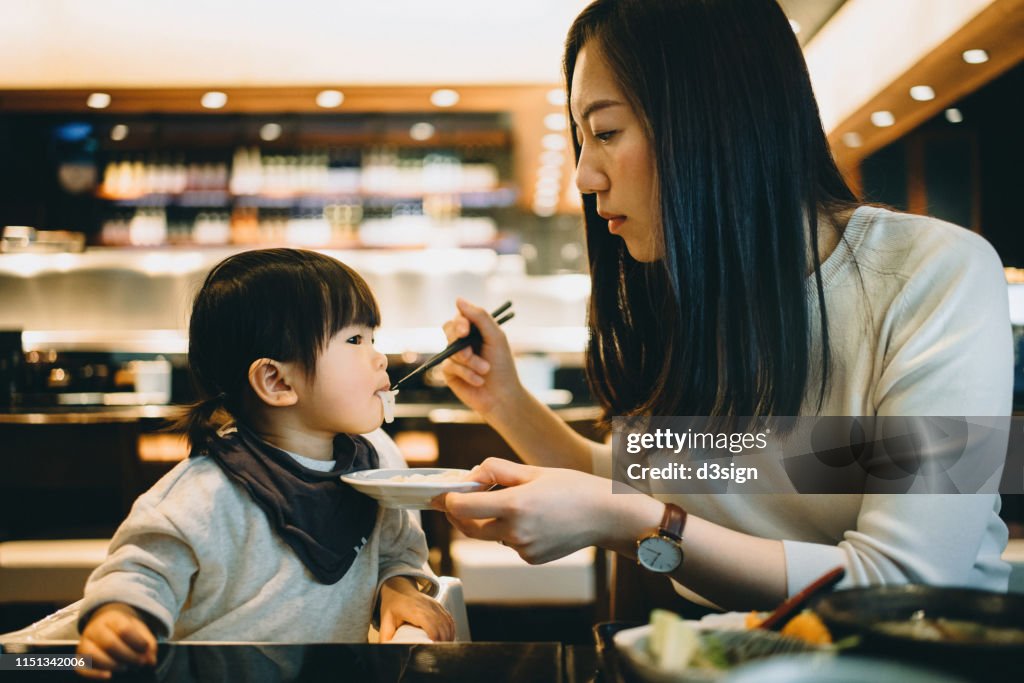Caring young mother feeding noodles to cute little daughter while having meal in the restaurant