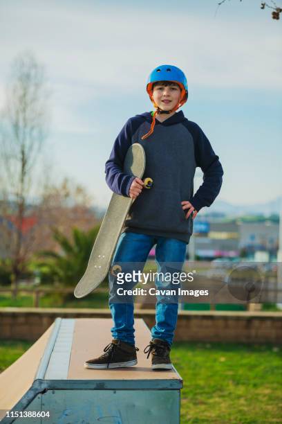 front view of young boy holding board while standing at skate ramp - boy skatepark stock-fotos und bilder