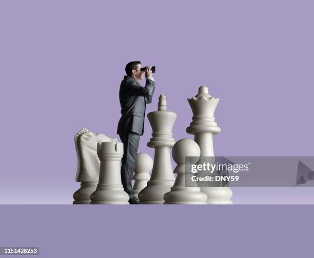 businessman looks through binoculars as he stands among chess pieces - spy glass businessman stock pictures, royalty-free photos & images