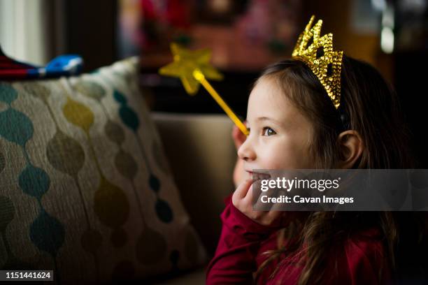 a smiling little girl in window light wears a golden crown and wand - princesa fotografías e imágenes de stock