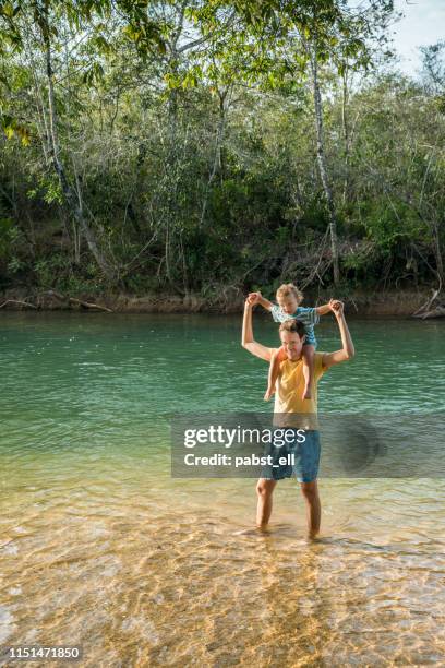 junger mann, der sohn auf seinen schultern trägt, steht im fluss _ - mato grosso do sul stock-fotos und bilder