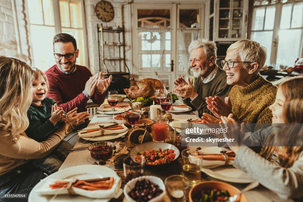 Happy extended family applauding during Thanksgiving meal at dining table.
