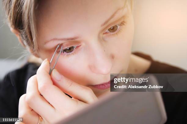 portrait of a european young girl who plucks eyebrows tweezers, looks in the mirror. close-up of a woman's face removing facial hair. eyebrow shaping and care. - tweezing eyebrows stock pictures, royalty-free photos & images