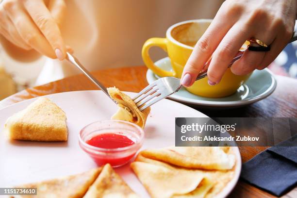 pancakes with berry jam on a white plate on a wooden background close-up. fork and knife in the hands of a woman. next is a mug of cappuccino. girl eating pancakes with cutlery in a cafe or restaurant - moitié photos et images de collection