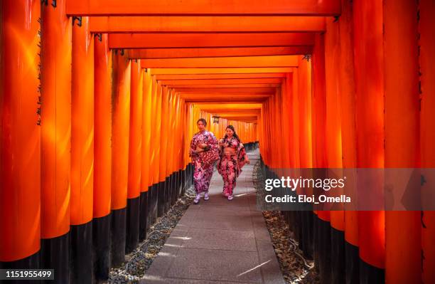 zwei japanerinnen, die durch die torii-tore bei fushimi inari taisha mit selfie-stock und kamera, kyoto, spazieren. japan - torii tor stock-fotos und bilder