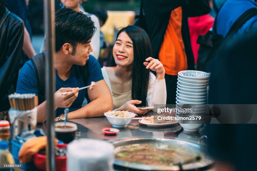 Encantadora pareja asiática joven disfrutando de la comida callejera en el mercado nocturno, Taipéi.